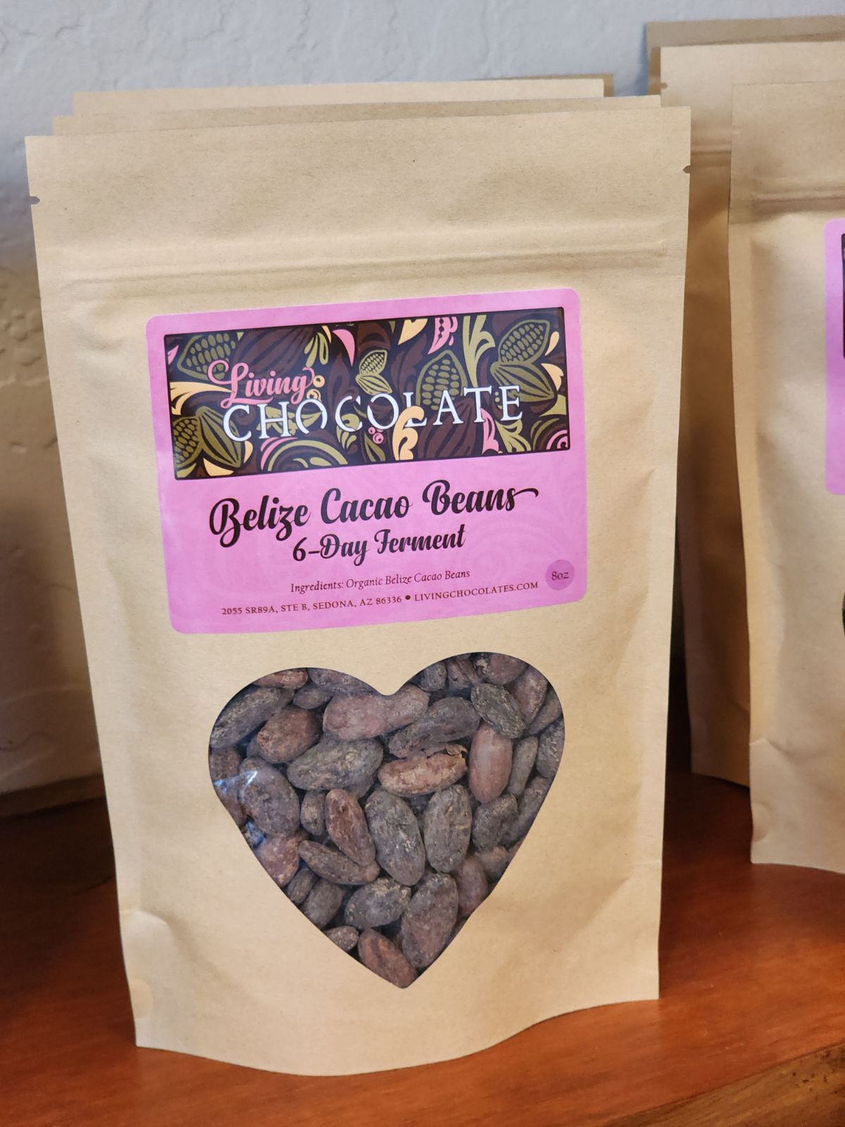 A light brown bag with a pink label filled with 6 day fermented Belize Cacao Beans with a heart shaped opening, sitting atop a wooden shelf.