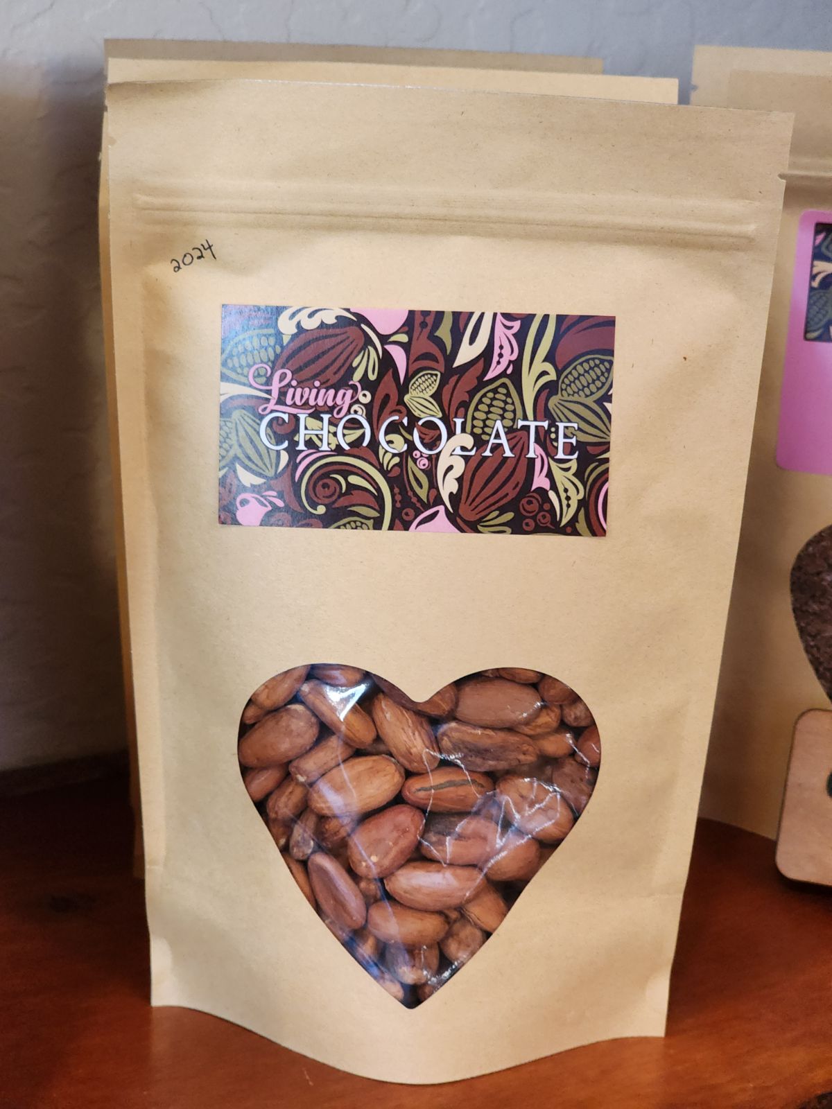 A light brown bag with Living Chcolate's logo, filled with Raw Cacao Beans from India, with a heart shaped opening, sitting atop a wooden shelf.